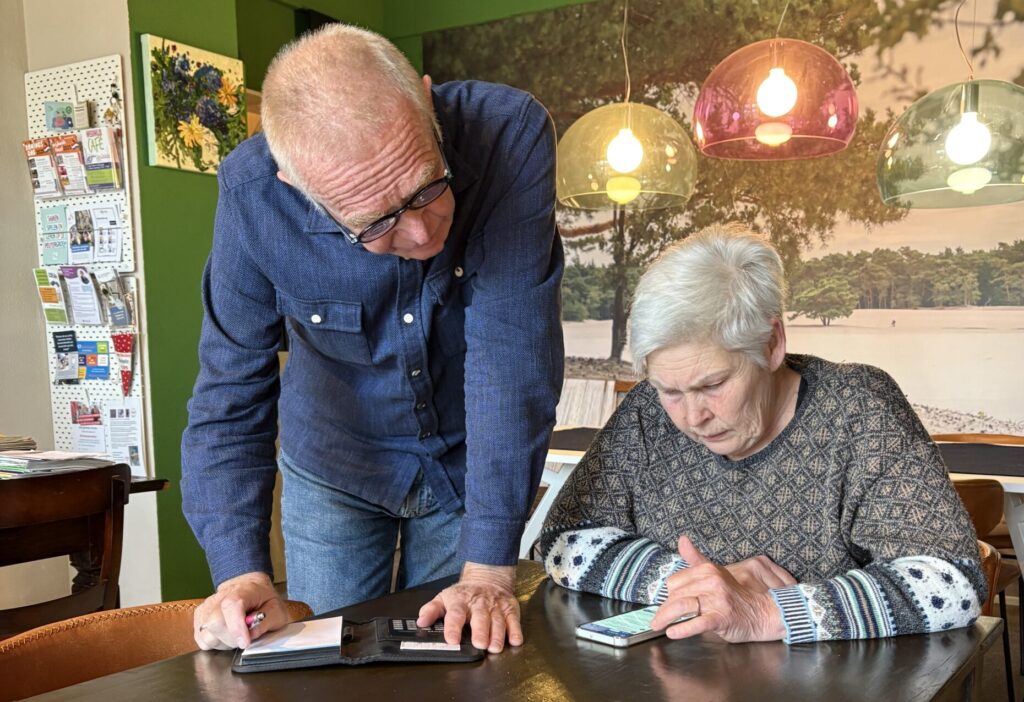 Meneer staat naast mevrouw die aan tafel zit. Ze kijken samen op de mobiele telefoon de voor hen op tafel ligt.